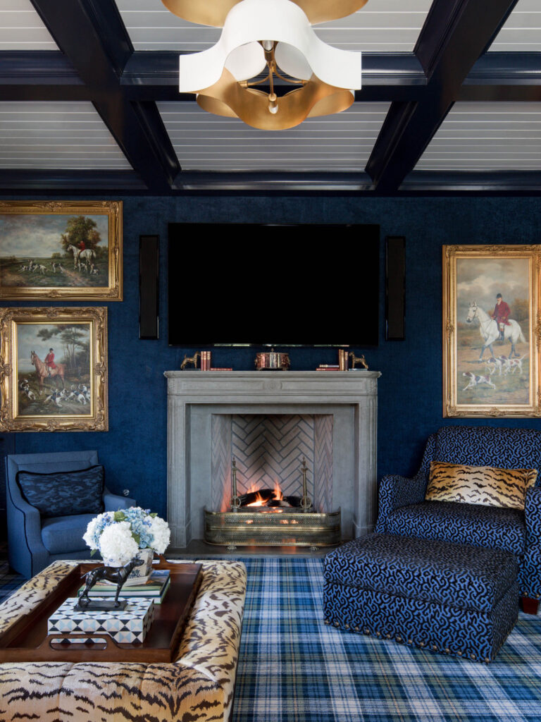 A cozy, elegant living room reflects the exuberant interior of this French Norman-inspired home, with a lit fireplace under a book-lined mantel, gold-framed equestrian paintings, blue patterned armchairs, and a tiger-print bench for a luxurious classic touch.