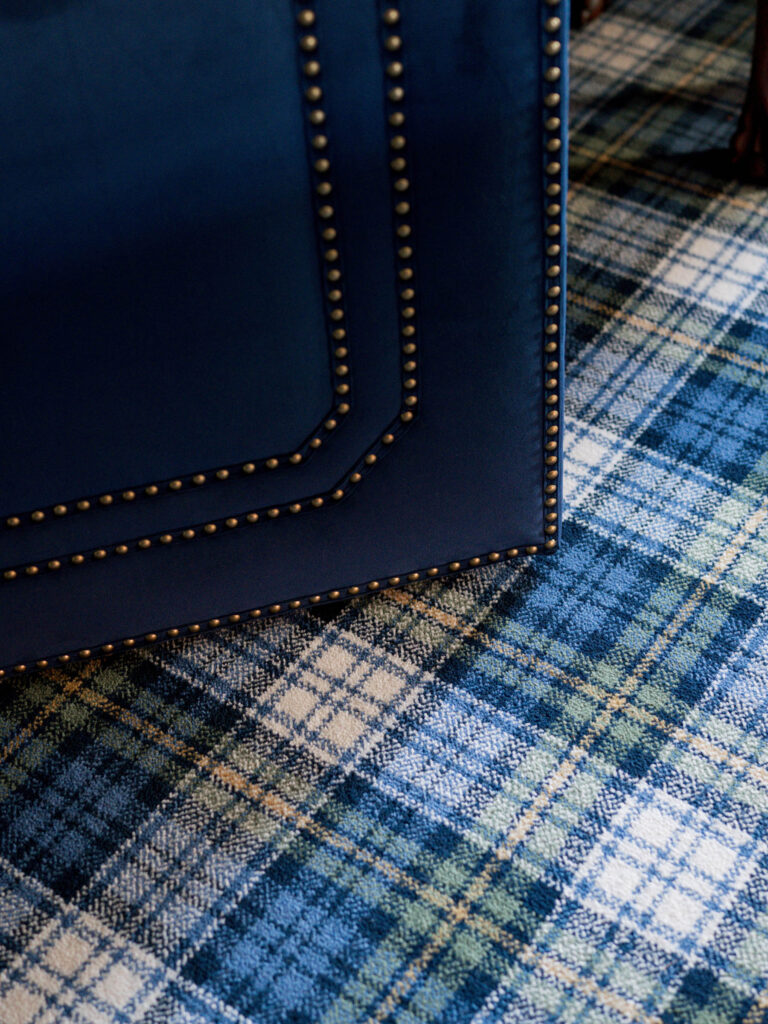 A close-up of a blue upholstered object with brass nailhead trim sits on a blue, grey, and white plaid-patterned carpet—details that hint at the exuberant interior of this French Norman-inspired home.