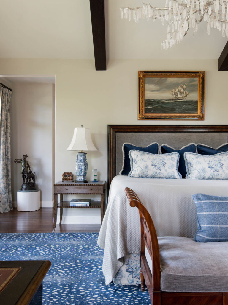 Elegant bedroom with a bed featuring blue and white pillows and bedding, a wooden bench, blue rug, side table with lamp, decorative statue, and framed ship painting above the bed; an exuberant interior of this French Norman-inspired home.