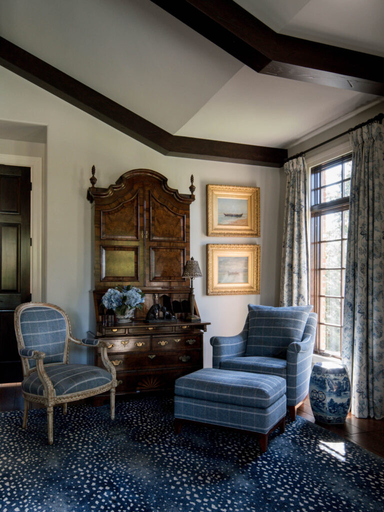 A cozy reading nook in the exuberant interior of this French Norman-inspired home features a blue plaid armchair and ottoman by a window, patterned drapes, an ornate wood armoire, framed paintings, and blue and white pottery accents.