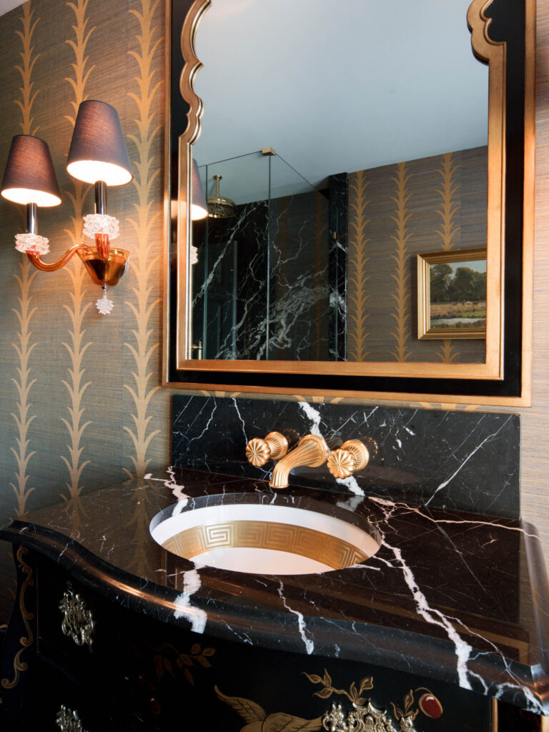 Luxurious bathroom in the exuberant interior of this French Norman-inspired home features a black marble vanity with white veining, gold fixtures, an oval Greek key sink, ornate gold wallpaper, a gold-framed mirror, and elegant brass lighting.