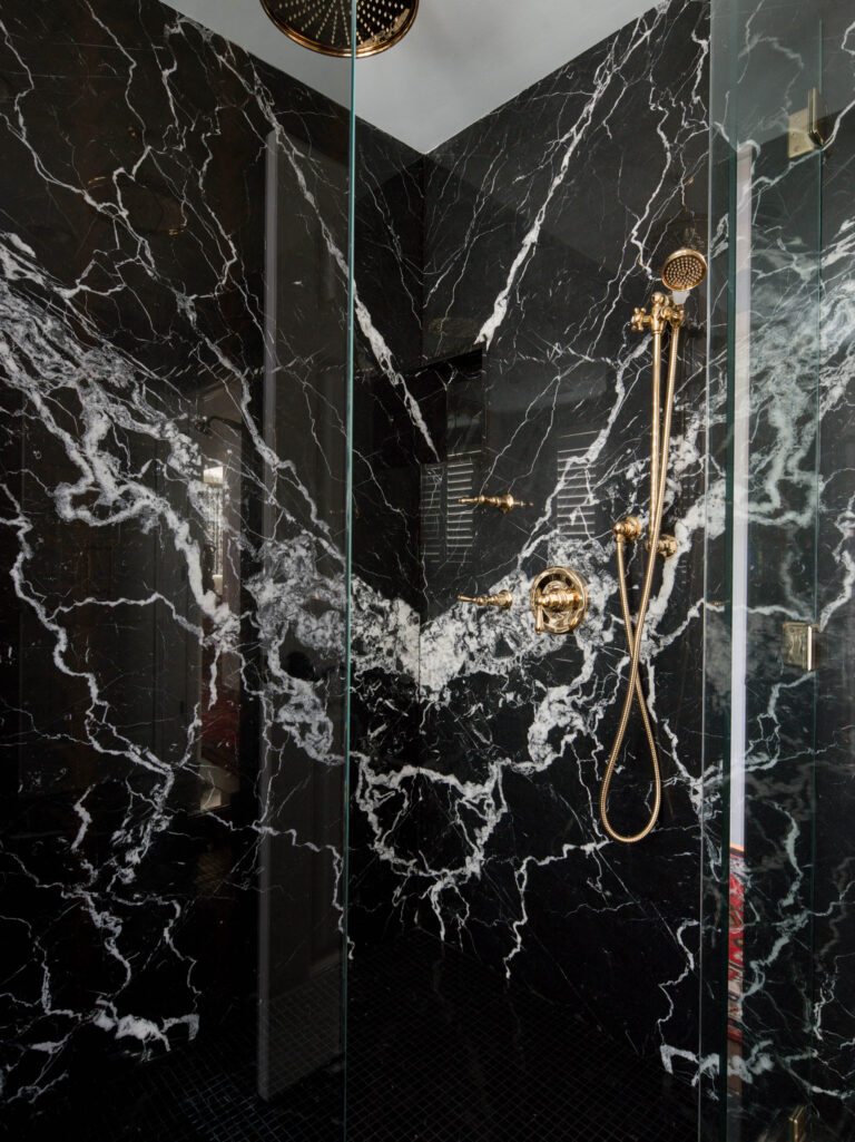 A modern shower with dramatic black marble walls, bold white veins, and gold fixtures defines the exuberant interior of this French Norman-inspired home. Subtle lighting and a glass door enhance its luxurious atmosphere.