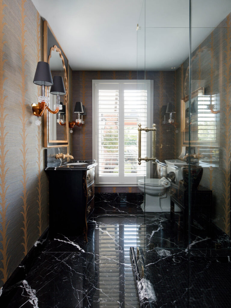 Elegant bathroom with black marble floor, gold and gray patterned wallpaper, black vanity with brass fixtures, and an ornate wall mirror—all echoing the exuberant interior of this French Norman-inspired home. Large window and white shutters invite abundant natural light.