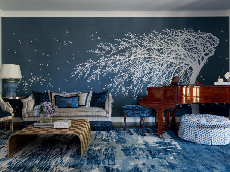 A stylish living room in the exuberant interior of this French Norman-inspired home features a dramatic navy wall mural with a wind-swept tree, velvet sofa, grand piano, modern coffee table, blue ottoman, and a blue-and-white rug.