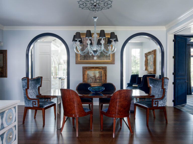 Elegant dining room with dark wood floors, a rectangular table, and six high-back velvet chairs. A glass chandelier hangs above. Two arched doorways hint at the exuberant interior of this French Norman-inspired home, filled with natural light.