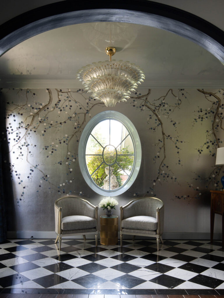 An elegant room in the exuberant interior of this French Norman-inspired home features a large oval window between two silver upholstered chairs, metallic floral walls, a crystal chandelier, black-and-white checkered tiles, and a flower-topped table.