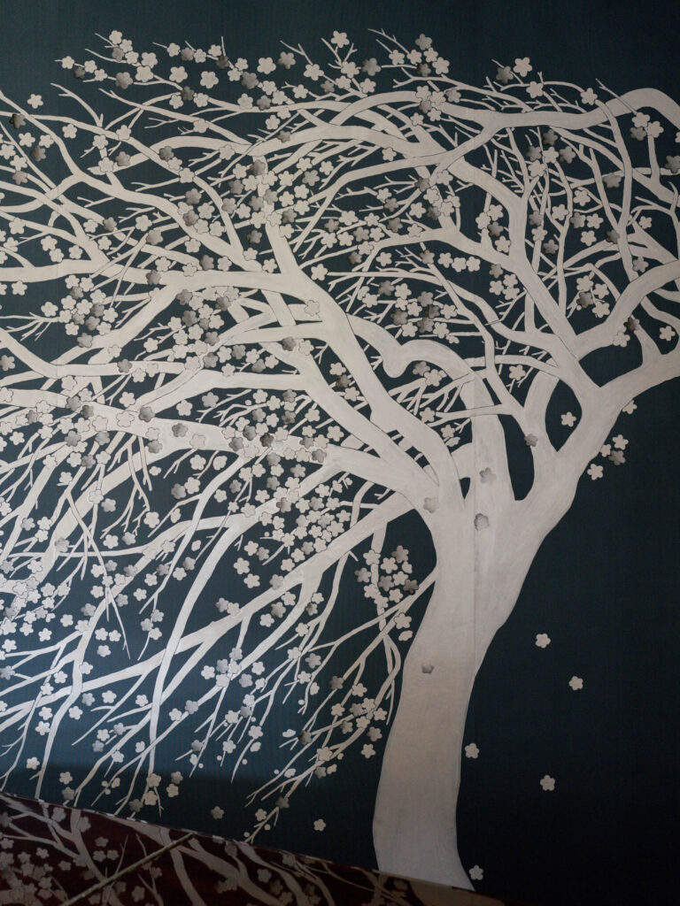A mural of a white cherry blossom tree with many branches and small flowers adorns the exuberant interior of this French Norman-inspired home, its blossoms scattered by the wind against a dark background, creating a dynamic sense of movement.