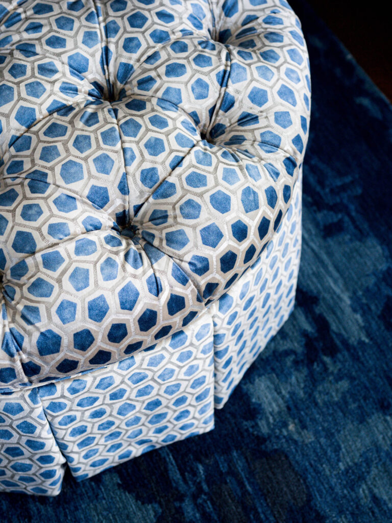 Close-up of a tufted ottoman upholstered with a geometric hexagon pattern in blue, gray, and white. Set on a deep blue rug, it showcases fabric details and plush design that enhance the exuberant interior of this French Norman-inspired home.