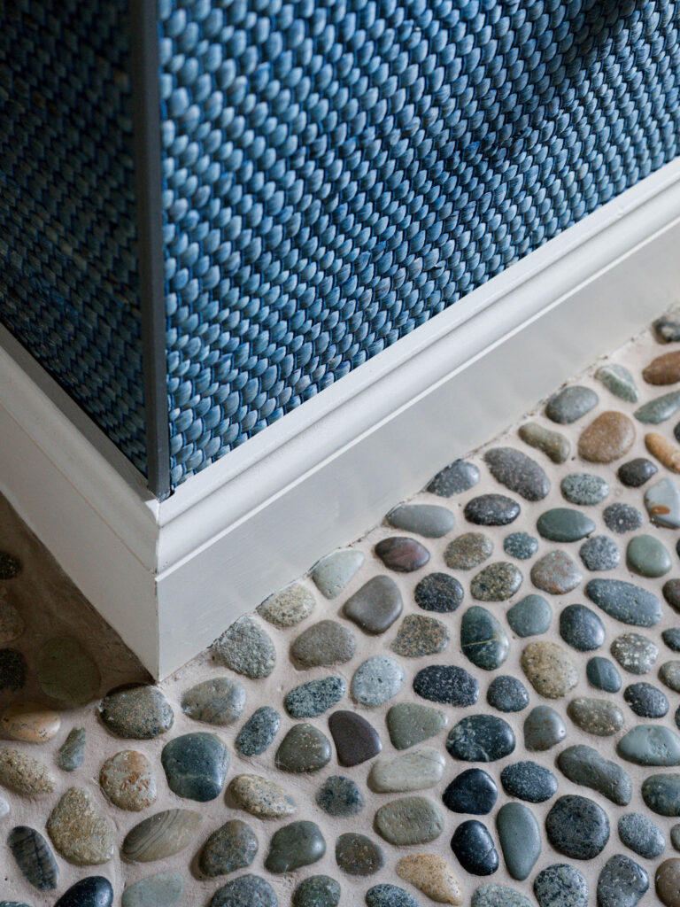 Close-up of a corner where a blue textured wall with a white baseboard meets a river stone floor, highlighting the exuberant interior of this French Norman-inspired home through contrasting textures and a natural, spa-like ambiance.